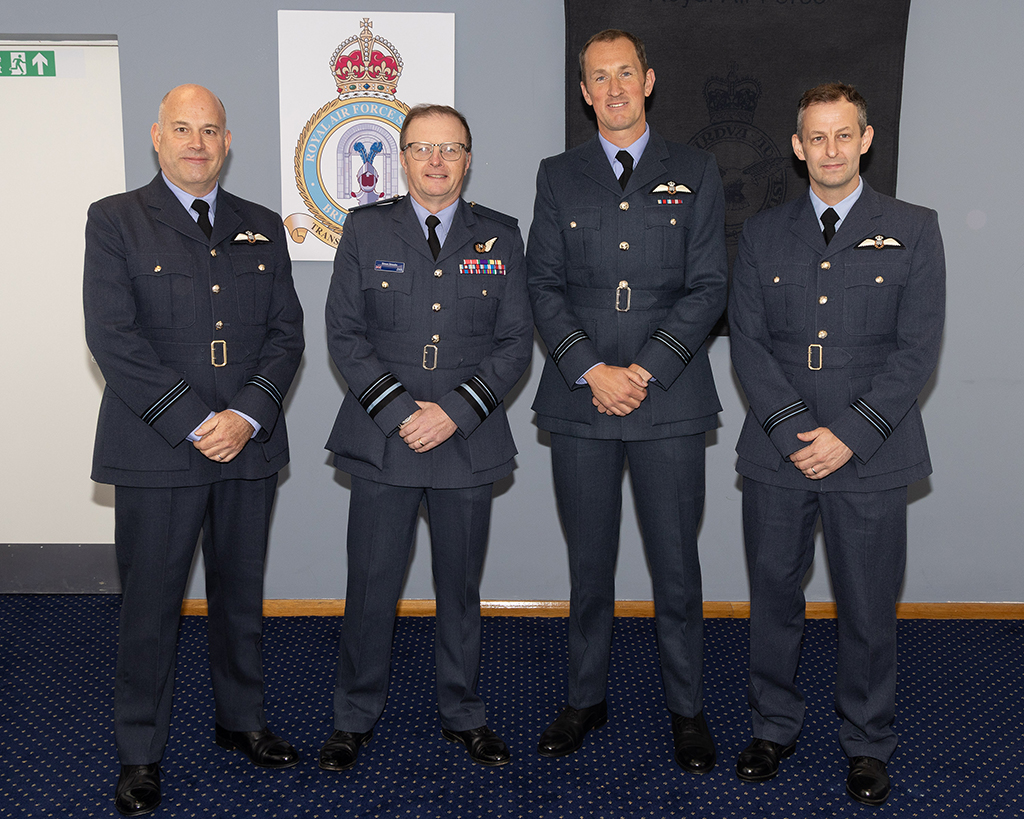 An official indoor photograph features, left, one 'RAF Wings' recipient, followed by Air Vice-Marshal Simon Strasdin, and then two 'RAF Wings' recipients as they stand in front of a grey wall decorated with two RAF signs.